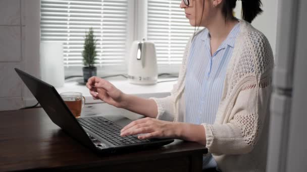 Freelancer, programador, programador, hacker, concepto de red social de comunicación. Mujer atractiva con gafas está en la cocina trabajando, mirando la pantalla del ordenador portátil y escribiendo algo en el teclado beber té — Vídeos de Stock