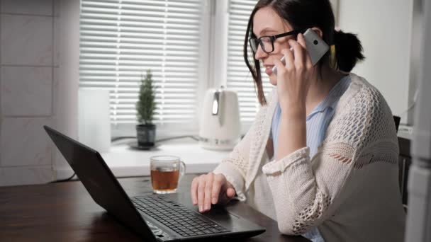 Chica freelancer en el concepto de trabajo. Mujer joven atractiva está trabajando en la cocina, hablando por teléfono en su mano izquierda, y con la derecha, se está concentrando en algo en el teclado del ordenador portátil — Vídeo de stock