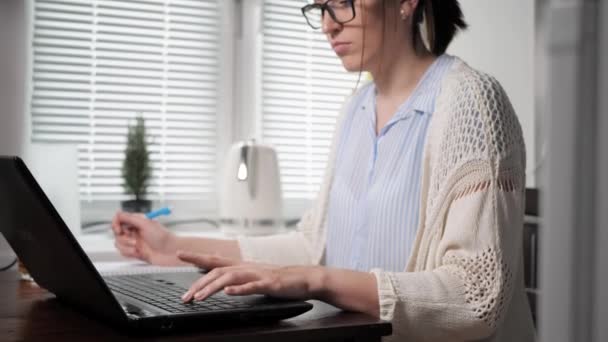 Freelancer Mädchen bei der Arbeit Konzept. Junge Frau mit attraktiver Brille arbeitet in der Küche mit Laptop, tippt mit der einen Hand etwas auf die Tastatur und schreibt mit der anderen Daten ins Notizbuch — Stockvideo