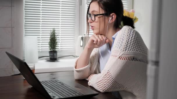 Frilansare flicka på jobbet koncept. Attraktiv ung kvinna i glasögon och med hörlurar tittar på laptop. Hon är i köket i lägenheten, pratar på videolänk med någon. Medelhög skott — Stockvideo