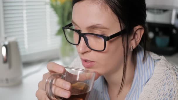 Menina bebe chá verde ou preto. Close-up de mulher jovem atraente com óculos na cozinha bebendo lentamente bebida marrom de caneca transparente e olhando em algum lugar pensativo com concentração — Vídeo de Stock