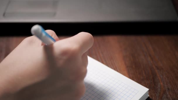 Main féminine écrit quelque chose dans un cahier avec stylo, à côté de lui est un ordinateur portable. Gros plan — Video