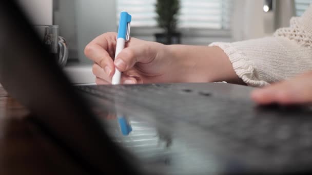 La mano femenina izquierda está escribiendo algo en el teclado del ordenador portátil, la mano derecha está escribiendo algo con bolígrafo en el portátil. Chica en el trabajo o concepto de estudio. Primer plano — Vídeos de Stock
