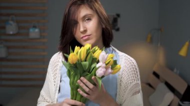 Çiçekli kız, elinde bir buket sarı laleyle kameraya bakan çekici genç kadın yüzüne çiçekler getiriyor ve güzel lalelerin tomurcuklarını kokluyor. Yavaş çekim ve yakın çekim