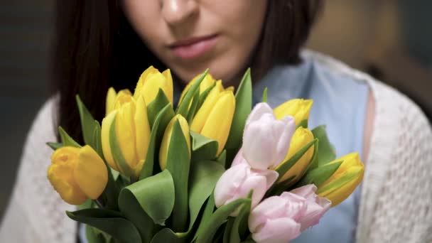 Chica con flores, mujer joven atractiva trae ramo de tulipanes amarillos a su cara y huele plantas para mirar a la cámara. La cámara se mueve hacia el modelo. cámara lenta y primer plano — Vídeo de stock