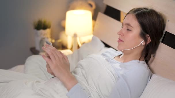 A menina ouve música e usa o telefone antes de ir para a cama. Jovem mulher atraente está deitada na cama sozinha no quarto, ela está ouvindo música com fones de ouvido e folheando através da tela celular. Close-up — Vídeo de Stock