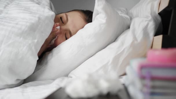Больная женщина насморк лежит в постели под одеялом и высморкается в бумажную салфетку. Холод, грипп, аллергия, пыль, кошачьи волосы, ринит, гипотермия, концепция сухого воздуха. Крупный план — стоковое видео