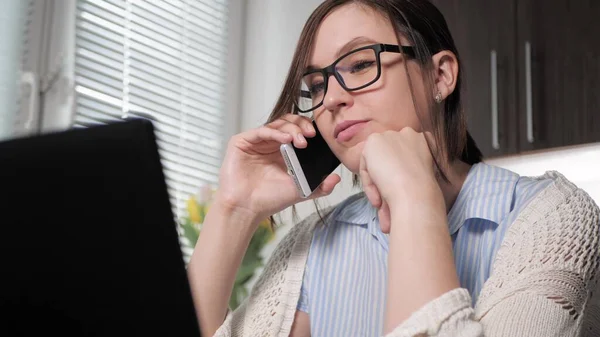 Freelancer girl at work. Attractive young woman in glasses talking on phone in kitchen, looking at laptop, agrees with something said on mobile. Close-up — Stock Photo, Image