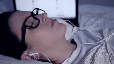 Kız gece müzik dinler. Gözlüklü ve kulaklıklı çekici genç bir kadın laptopun yanında yatar ve müzik dinlemekten hoşlanır. Yakın plan.