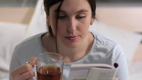 La chica está leyendo un libro. Joven mujer caucásica atractiva se sienta en la cama por la noche y lee el libro y bebe té negro antes de acostarse. Primer plano — Foto de Stock