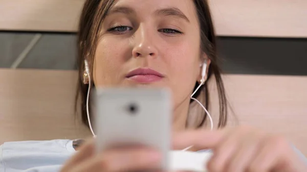 Girl listens to music and uses phone. Young attractive woman is lying in bed with headphones listening to music and leafing through her cell screen. Close-up