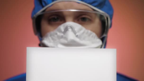 Θολή κοπέλα με μάσκα αναπνευστήρα μπλε προστατευτική στολή και γυαλιά κρατά λευκό σημάδι στα χέρια της και γνέφει το κεφάλι της με την έγκριση. Μόλυνση, προειδοποίηση, επιδημία, πανδημία, COVID-19 έννοια. Κλείσε. — Αρχείο Βίντεο