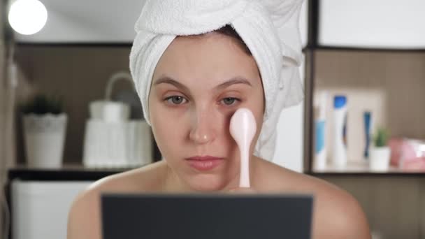 Chica utiliza masajeador para los párpados inferiores. Mujer atractiva en el baño se ve en el espejo y utiliza masaje de la piel cerca de sus ojos. Higiene, esteticista, maquillaje, concepto de cuidado facial. Primer plano — Vídeo de stock