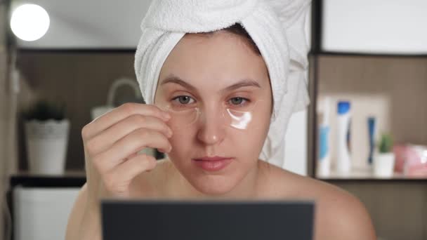 Meisje in de badkamer met handdoek op haar hoofd kijkt in de spiegel en verwijdert ooglapjes. Hygiëne, schoonheidsspecialiste, make-up, gezichtsverzorging. Close-up — Stockvideo