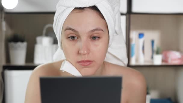 Дівчина потирає обличчя. Приваблива жінка у ванній з білим рушником на волоссі дивиться у дзеркало і протирає обличчя мокрим рушником. Гігієна, косметолог, макіяж, концепція догляду за обличчям. Крупним планом — стокове відео