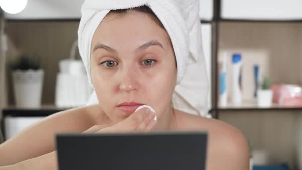 Girl rubs her face. Attractive woman in bathroom with white towel on her hair looks in mirror and rubs her face with cotton pad. Hygiene, beautician, makeup, face care concept. Close-up — Stock Video