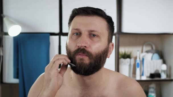 Чоловік розчісує свою бороду. Привабливий чоловік у ванній виглядає у дзеркалі і поєднує чорне волосся на обличчі. Гігієна, догляд за волоссям, концепція свіжості. Крупним планом — стокове відео