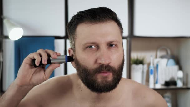 Mann schneidet sich Haare im Ohr. Junge attraktive Kerl mittleren Alters im Badezimmer schaut in den Spiegel und verwendet Trimmer, um seine Ohren zu schneiden. Hygiene, Frischekonzept. Nahaufnahme — Stockvideo