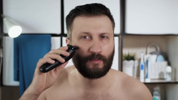 L'homme redresse ses cheveux sur la barbe. Homme attrayant dans la salle de bain regarde dans le miroir et coupe ses cheveux avec tondeuse faire bords de barbe même. Beauté, concept de fraîcheur. Gros plan — Video