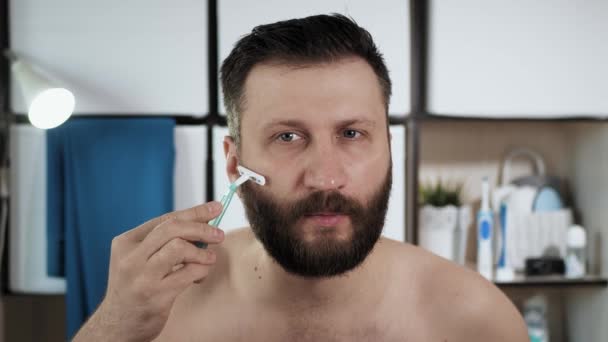 O homem endireita o cabelo na barba. cara caucasiano atraente no banheiro olha no espelho e rapa o cabelo com lâmina de barbear descartável fazendo bordas de barbas mesmo. Beleza, conceito de frescura. Close-up — Vídeo de Stock