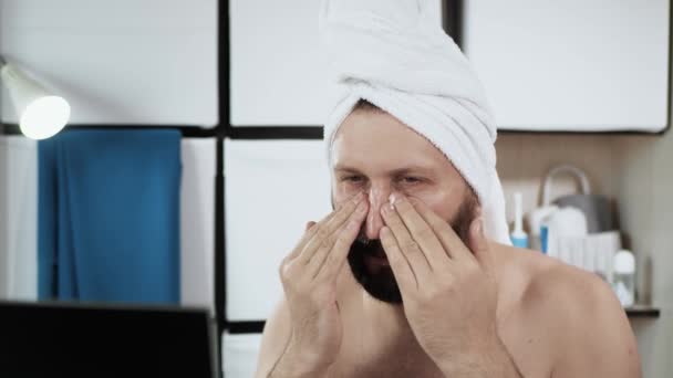 Man applies cream on his face. Travesty, bearded caucasian man with towel on his head applies wrinkle cream moisturizing his face. Close-up — Stock Video