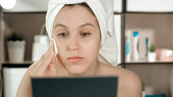 Girl squeezes black dots on her nose. Attractive woman in towel on her head looks in mirror and presses on skin on bridge of her nose, squeezing black dots. Hygiene, beautician, makeup, face care concept. Close-up view