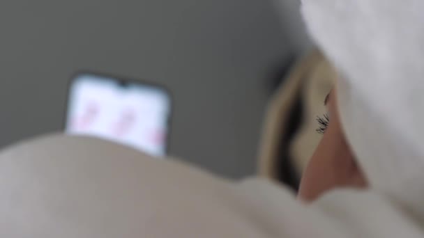Het meisje gebruikt internet via de telefoon. Vrouw met handdoek op haar hoofd en witte jas ligt op bed en gebruikt smartphone om nieuwsfeed door te bladeren. Close-up — Stockvideo