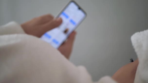 여자는 전화로 인터넷을 사용 한다. 머리에 타월을 쓰고 흰색 코트를 입은 여성은 침대에 누워 소식을 전하기 위해 스마트폰을 사용 한다. 클로즈업 — 비디오