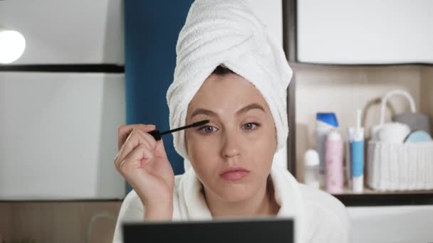 Meisje schildert wimpers. Jonge aantrekkelijke vrouw in witte handdoek en badjas kijkt in de badkamer spiegel en schildert wimpers met mascara. Schoonheidsspecialiste, make-up alleen thuis concept. Close-up — Stockvideo