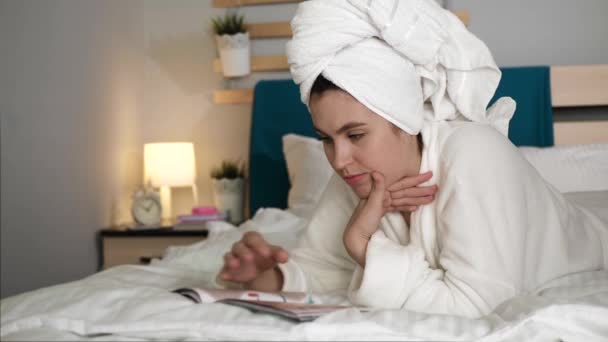 Девушка читает журнал. Женщина в спальне с полотенцем на голове и в белом халате лежит на животе в постели и переворачивает страницы модного журнала. Средний выстрел — стоковое видео