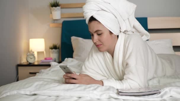 Girl writes message on phone. Woman in bedroom with towel on her head and in white coat lies on stomach in bed and dials message on touch screen of cell. Medium shot — Stock Video