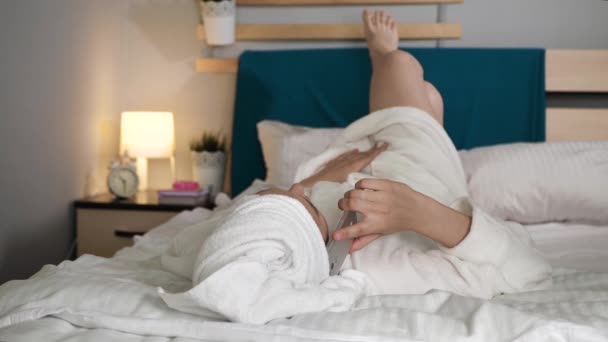 Meisje aan de telefoon. Vrouw in de slaapkamer met handdoek op haar hoofd en in witte badjas ligt op de rug en praat op haar mobiele telefoon. Middelmatig schot — Stockvideo