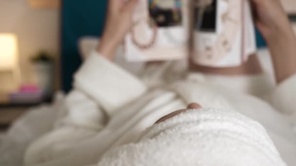 Revista de lectura de chicas. Visión borrosa de la mujer en el dormitorio en albornoz blanco se encuentra en su espalda y da la vuelta a las páginas de la revista de moda. Primer plano — Vídeo de stock