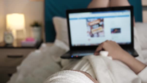 Meisje leest nieuws op laptop. Wazig zicht op vrouw in slaapkamer in witte badjas ligt op haar rug en scrollt pagina 's van het sociale netwerk op laptop. Close-up — Stockvideo