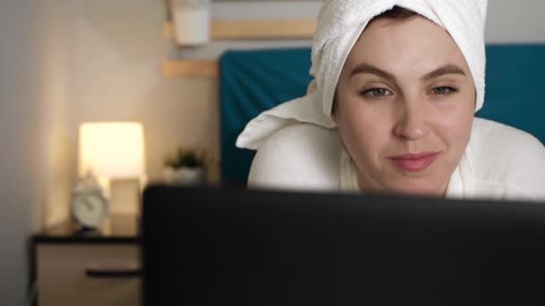 Девушка работает на ноутбуке. Привлекательная женщина в спальне в белом халате лежит на животе и пользуется ноутбуком. Крупный план — стоковое видео