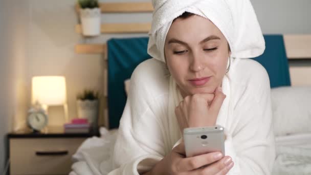 Дівчина прокручує телефон. Приваблива жінка в спальні в білому халаті з рушником на голові лежить на животі і гойдається пальцем на сенсорному екрані мобільного телефону. Крупним планом — стокове відео