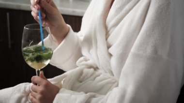 Kız mojito içiyor. Kafasında havluyla ve bornozlu kadın alkollü mojito içer ve pencereden dışarı bakar. Yakın plan.
