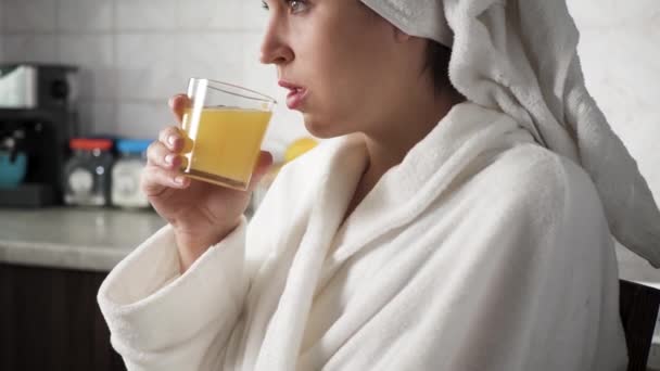 La chica está bebiendo jugo de naranja. Mujer en la cocina en albornoz blanco con toalla en la cabeza se sienta frente a la ventana y bebe jugo de naranja. Primer plano — Vídeo de stock