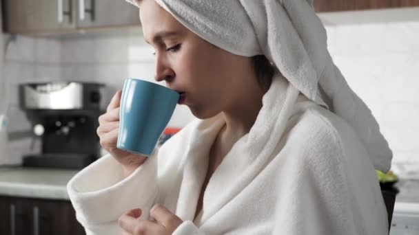 A rapariga está a beber café. Mulher na cozinha em roupão branco com toalha na cabeça senta-se na frente da janela e bebe café quente. Close-up — Vídeo de Stock