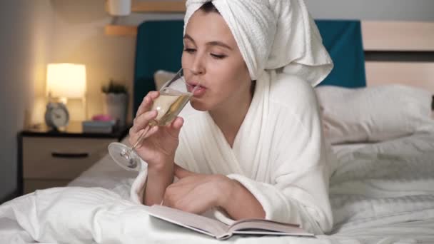 Şampanya içip kitap okuyan bir kız. Yatak odasındaki beyaz bornozlu çekici kadın kafasında havluyla yatakta uzanıyor şarap içiyor ve kitap okuyor, sayfaları çeviriyor. Yakın plan. — Stok video