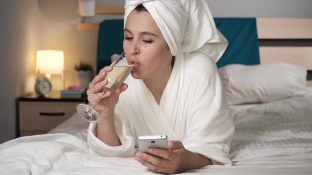 Девушка пьет вино и пользуется телефоном. Привлекательная женщина в спальне в белом халате с головой полотенца лежит в кровати, пьет шампанское и вводит текст на экранной клавиатуре сотового сенсорного экрана. Крупный план — стоковое видео