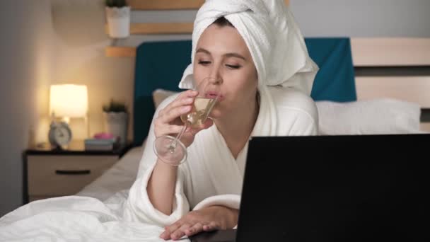 Girl drinking wine and working on laptop. Attractive woman in bedroom in white bathrobe lies in bed drinks champagne and uses laptop. Close-up — Stock Video