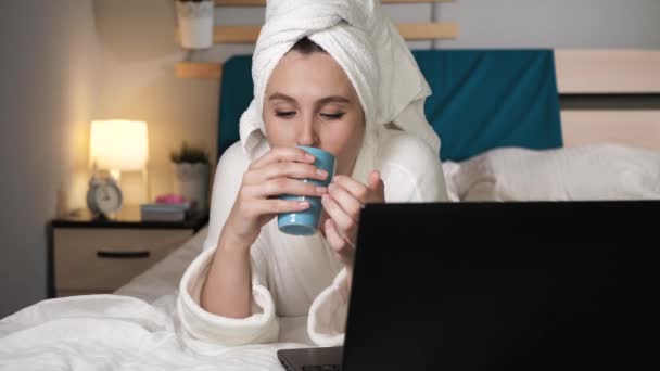 Chica bebiendo café o té y trabajando en el portátil. Mujer atractiva en el dormitorio en albornoz blanco se encuentra en la cama bebidas café caliente o té y utiliza el ordenador portátil. Primer plano — Vídeos de Stock