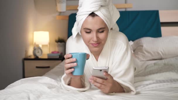 Chica bebiendo café o té y usar el teléfono. Mujer atractiva en el dormitorio en albornoz blanco se encuentra en la cama bebe café caliente o té e introduce texto en el teclado en pantalla de la pantalla táctil celular. Primer plano — Vídeos de Stock