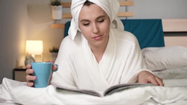 Chica bebiendo café o té y leyendo revista. Mujer atractiva en el dormitorio en albornoz blanco se encuentra en la cama bebiendo café caliente o té y da vuelta las páginas de la revista de moda. Primer plano — Vídeos de Stock