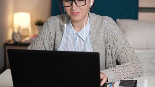 Flickan lyssnar på musik och jobbar vid datorn. Ung kvinna med glasögon ensam i sovrummet sitter vid skrivbordet lyssna på musik på hörlurar och skriva på laptop tangentbord. Arbete hemma, frilansande koncept — Stockvideo