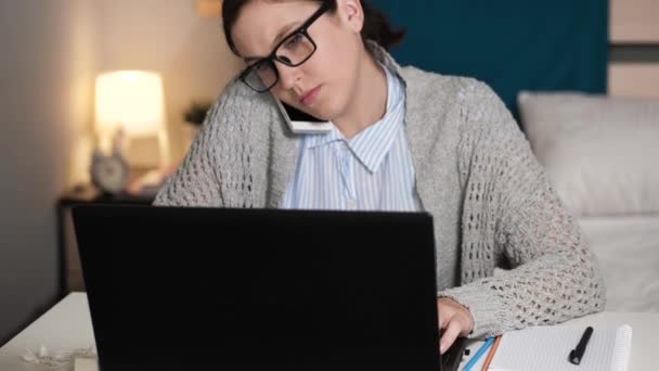 La chica está hablando por teléfono y trabajando en la computadora. Mujer atractiva con gafas solo en el dormitorio se sienta en el escritorio escribiendo en el teclado del ordenador portátil y hablando en el teléfono celular. Trabajo en casa, concepto freelance — Vídeos de Stock