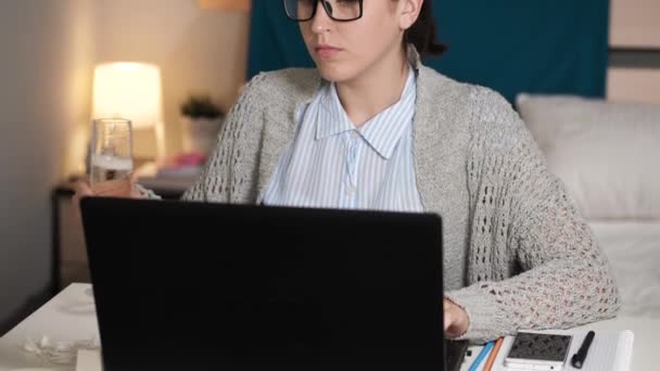 Flickan dricker vin och jobbar vid datorn. Kvinna med glasögon ensam i sovrummet sitter vid skrivbordet skriva på laptop tangentbord och dricka champagne. Arbete hemma, frilansande koncept — Stockvideo