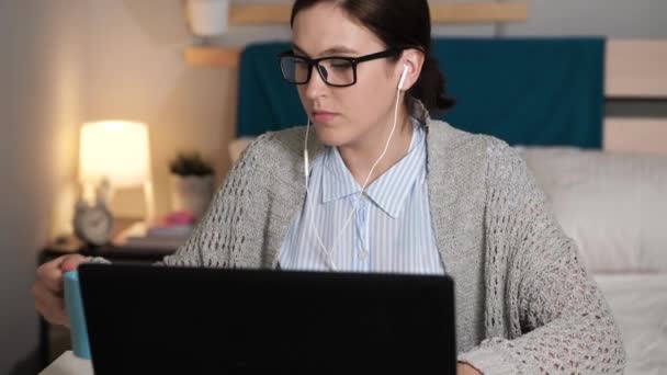 Dziewczyna słucha muzyki pije kawę lub herbatę i pracuje w komputerze. Kobieta w słuchawkach sama w sypialni siedzi przy biurku pisząc na klawiaturze laptopa i pijąc kawę lub herbatę. Praca w domu jako wolny strzelec — Wideo stockowe