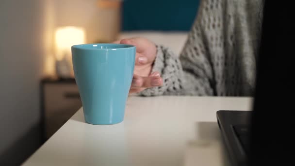 Mano femminile prende una tazza di caffè o tè dal tavolo e poi lo rimette a posto. La donna si siede alla scrivania a lavorare sul computer portatile e beve bevande calde. Lavoro a casa, concetto di freelance — Video Stock